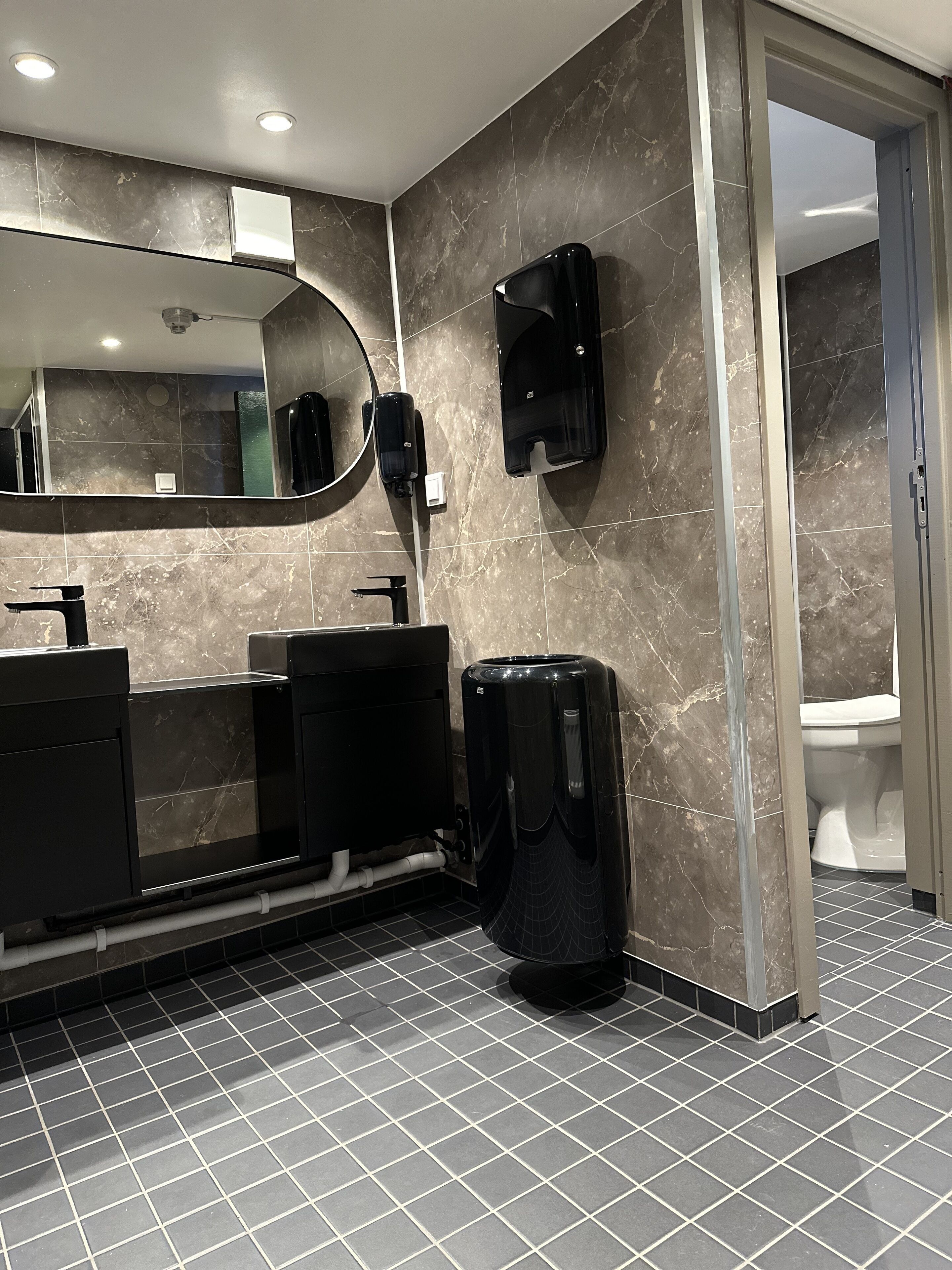 Une salle de bain moderne avec un grand miroir, deux lavabos, une poubelle noire et des toilettes visibles à travers une porte ouverte.