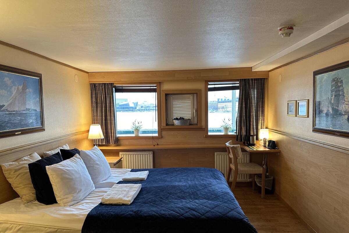 A hotel room with a large bed, two windows with curtains, a desk with a chair, and a radiator.
