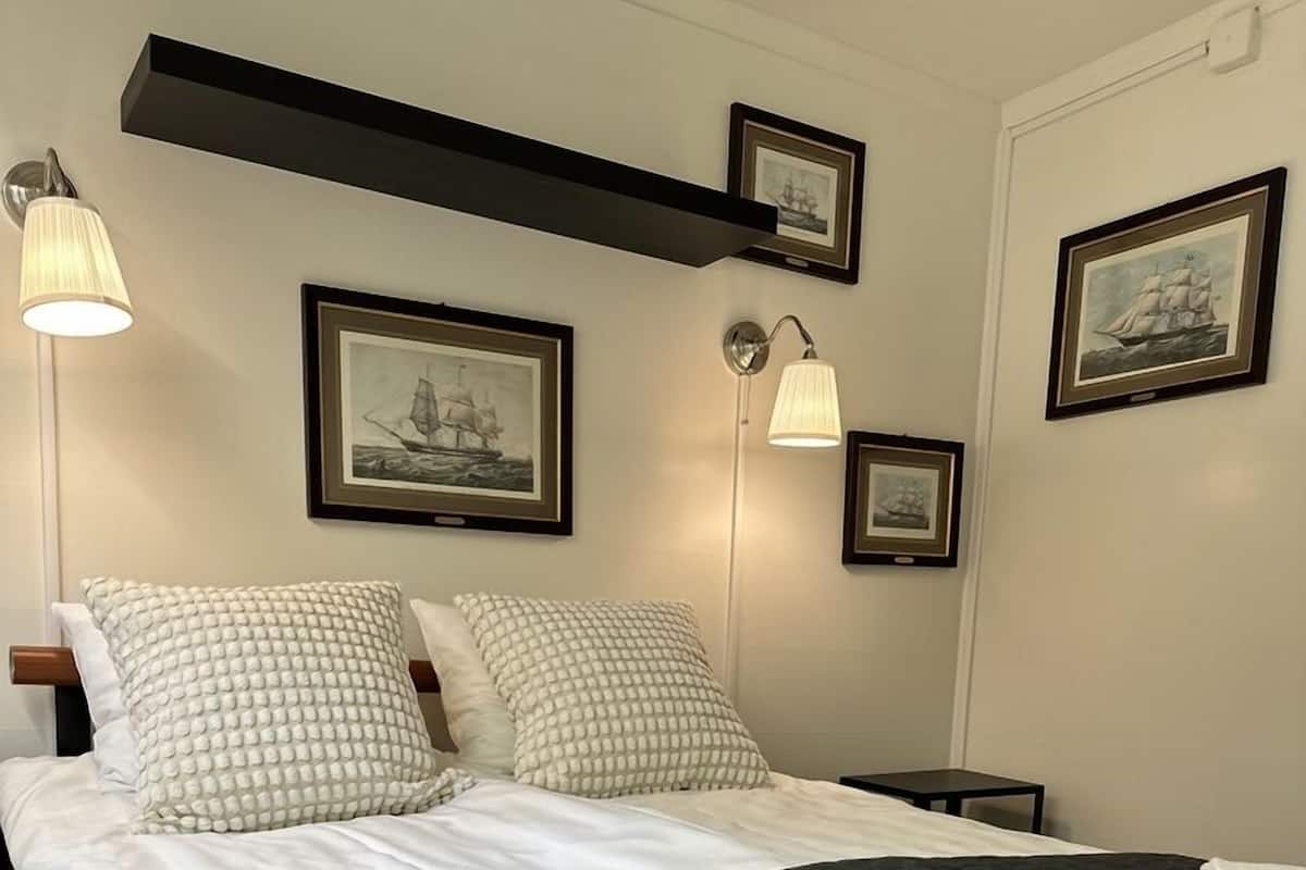 A neatly made bed with a dark quilt, two pillows, and a folded towel on a black headboard. There are two bedside tables, one with a lamp and another with a framed picture.