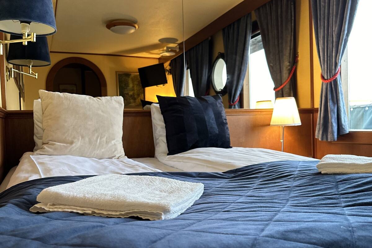 A neatly made bed with a blue quilt and white pillows, a bedside lamp, and a window with curtains.
