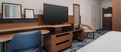 A hotel room with a bed, a desk with a flat-screen TV, a chair, a dresser, and a mirror.