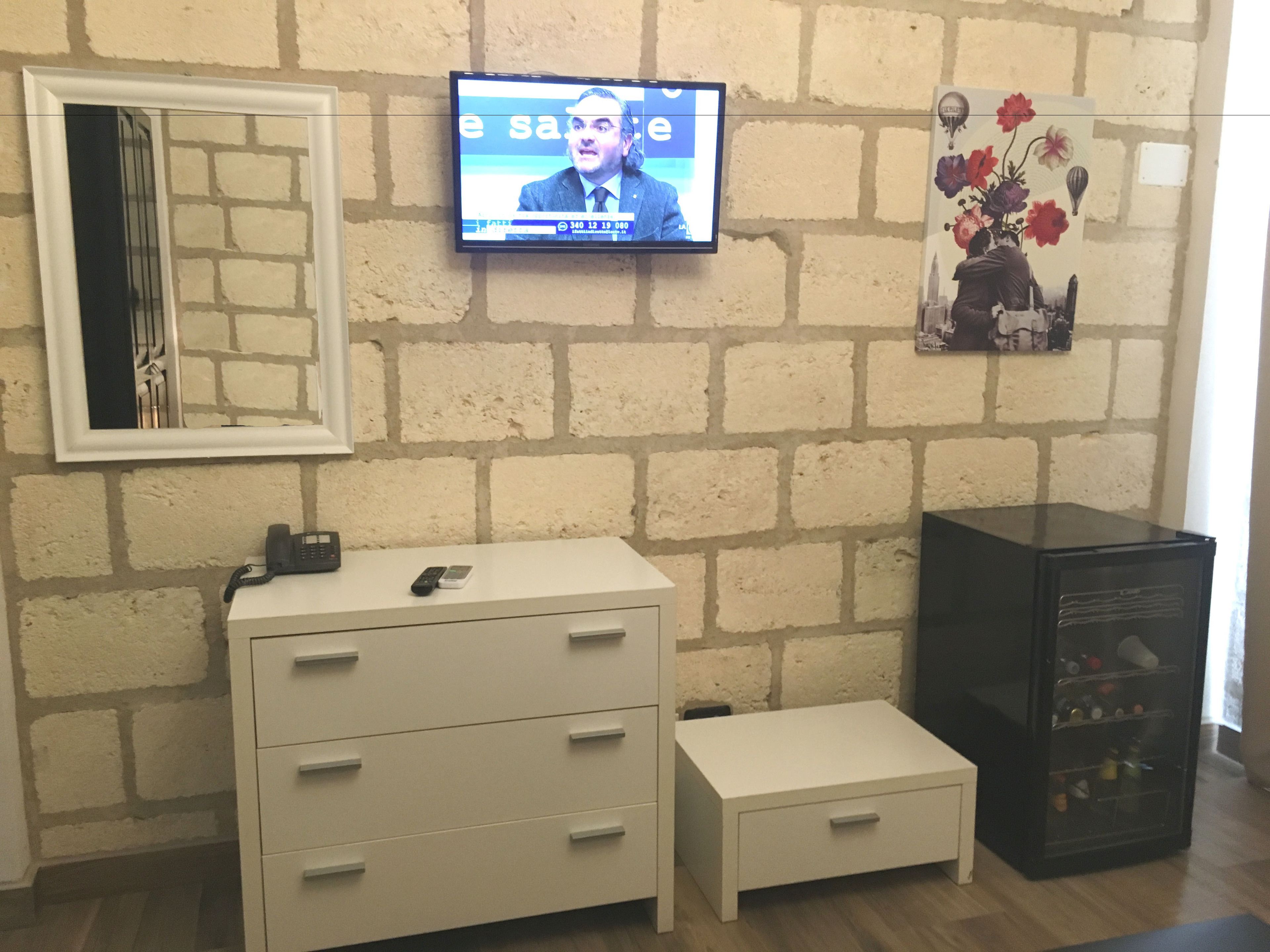 A room with a brick wall, a flat-screen TV mounted above a white dresser with drawers, a small white nightstand, and a black minibar.