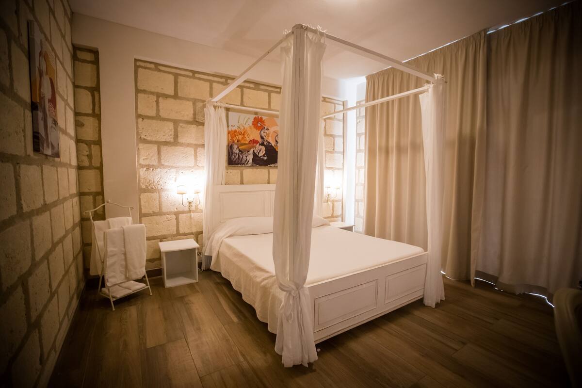 A bedroom with a four-poster bed, a nightstand, a wall-mounted painting, and a brick wall.