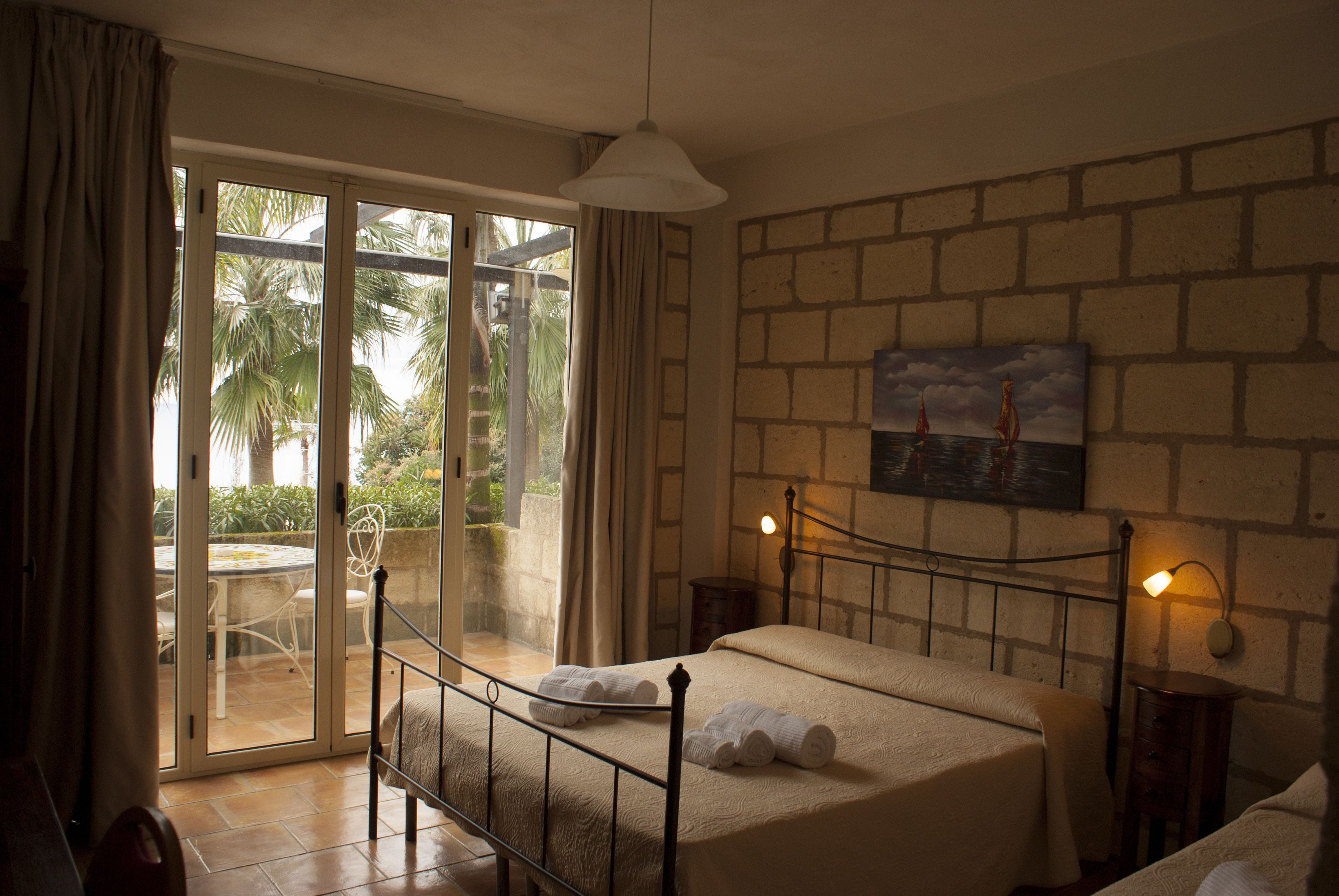 A bedroom with a bed, a nightstand, a lamp, a painting, and a sliding glass door leading to a patio with a table and chairs.