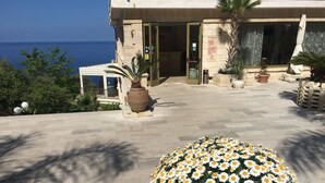 Ein Gebäude mit einer Steinfassade, einer großen Blumenkomposition im Vordergrund und Blick auf das Meer im Hintergrund.