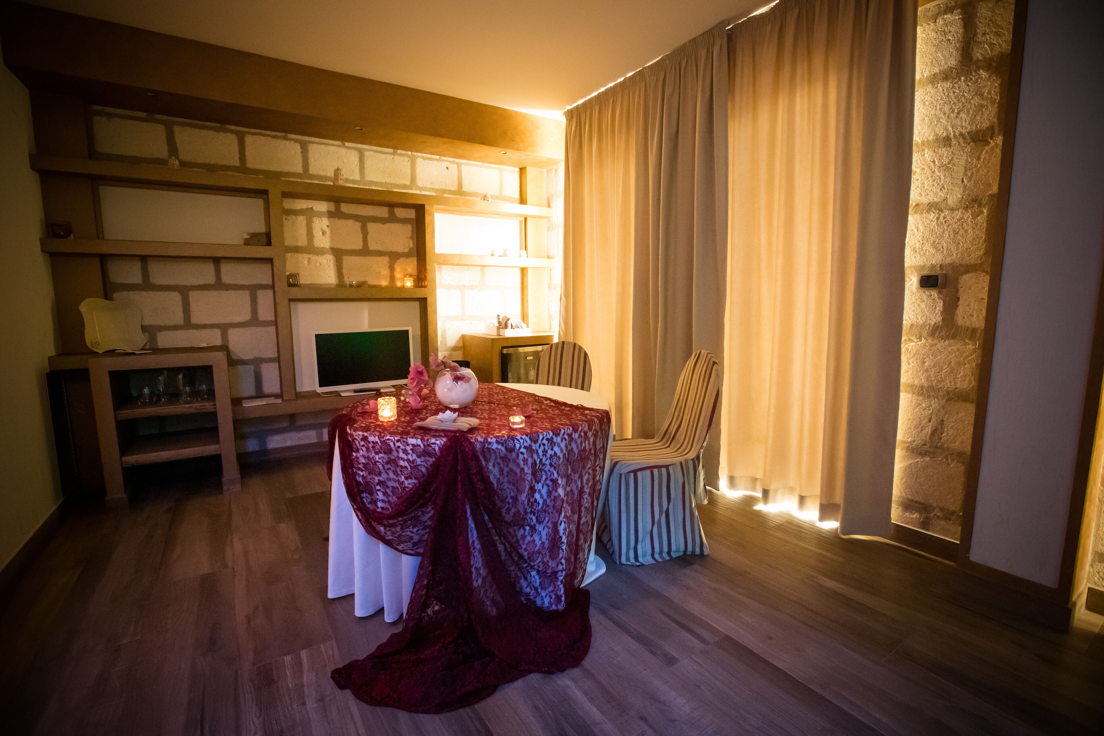A cozy room with a wooden floor, a table with a red tablecloth, a television, and a stone wall.