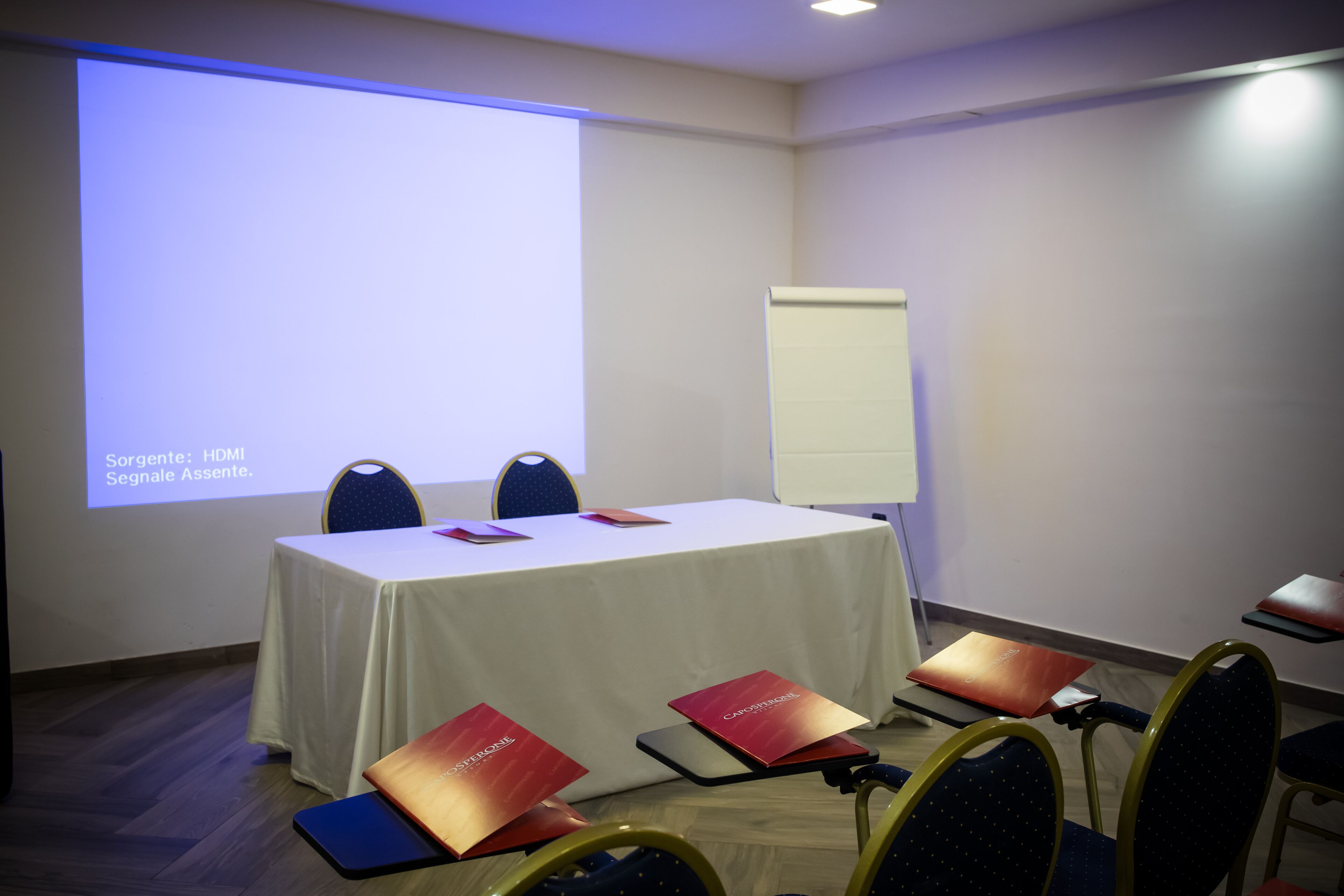 Uma sala de conferência com uma mesa branca, quatro cadeiras e uma tela de projetor exibindo texto.