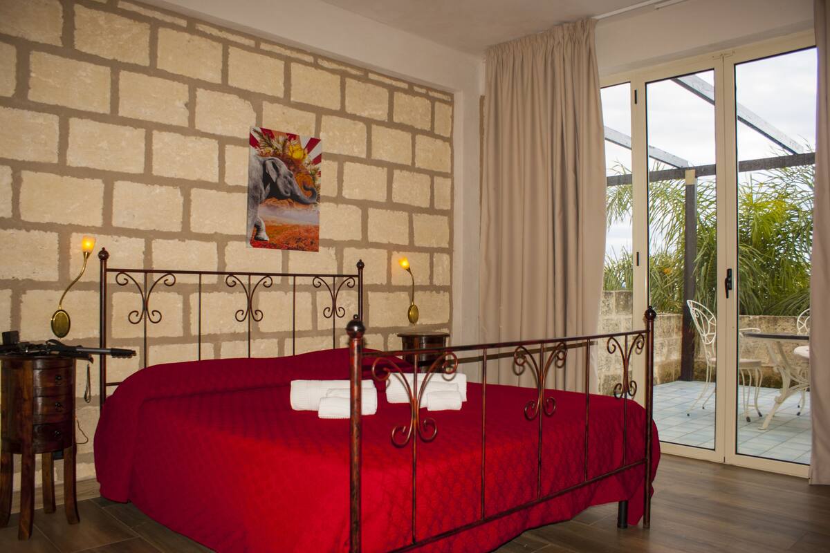 A bedroom with a red bedspread, a brick-patterned wall, and a view of an outdoor patio.