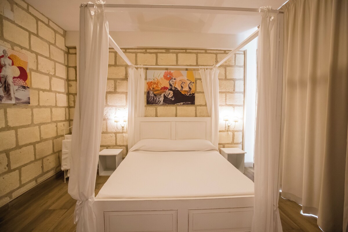 A room with a four-poster bed, white curtains, a brick wall, and a painting above the bed.
