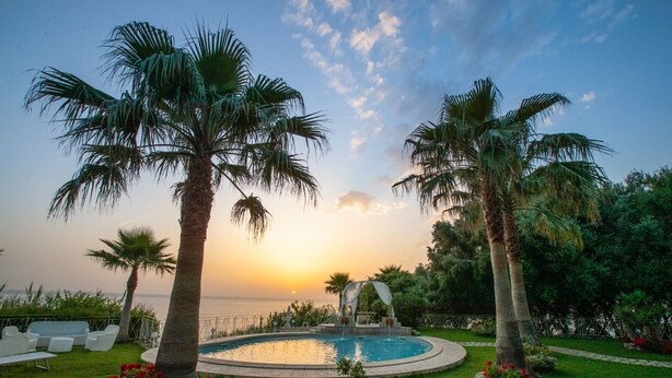 Ein Poolbereich mit Palmen, einem runden Pool und Blick auf das Meer bei Sonnenuntergang.
