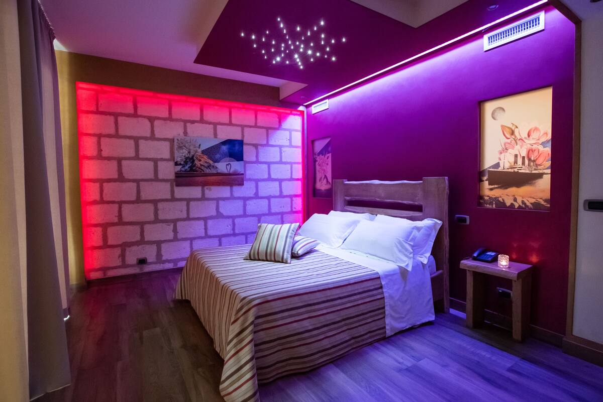 A bedroom with a brick-patterned wall, a bed with striped bedding, a nightstand with a lamp, and a ceiling light fixture.