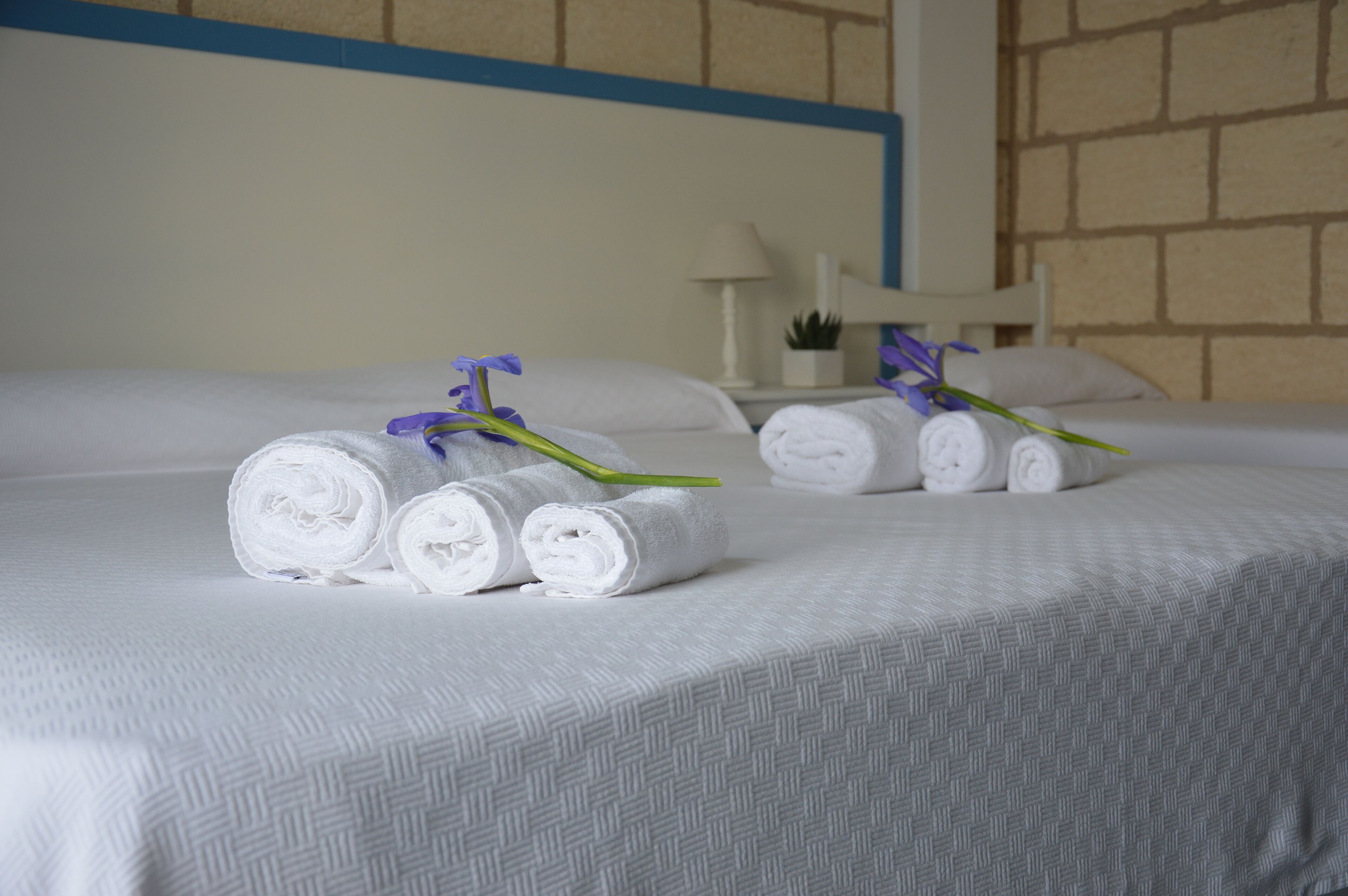 A neatly made bed with rolled white towels and a purple flower, a white nightstand with a lamp, and a brick-patterned wall.