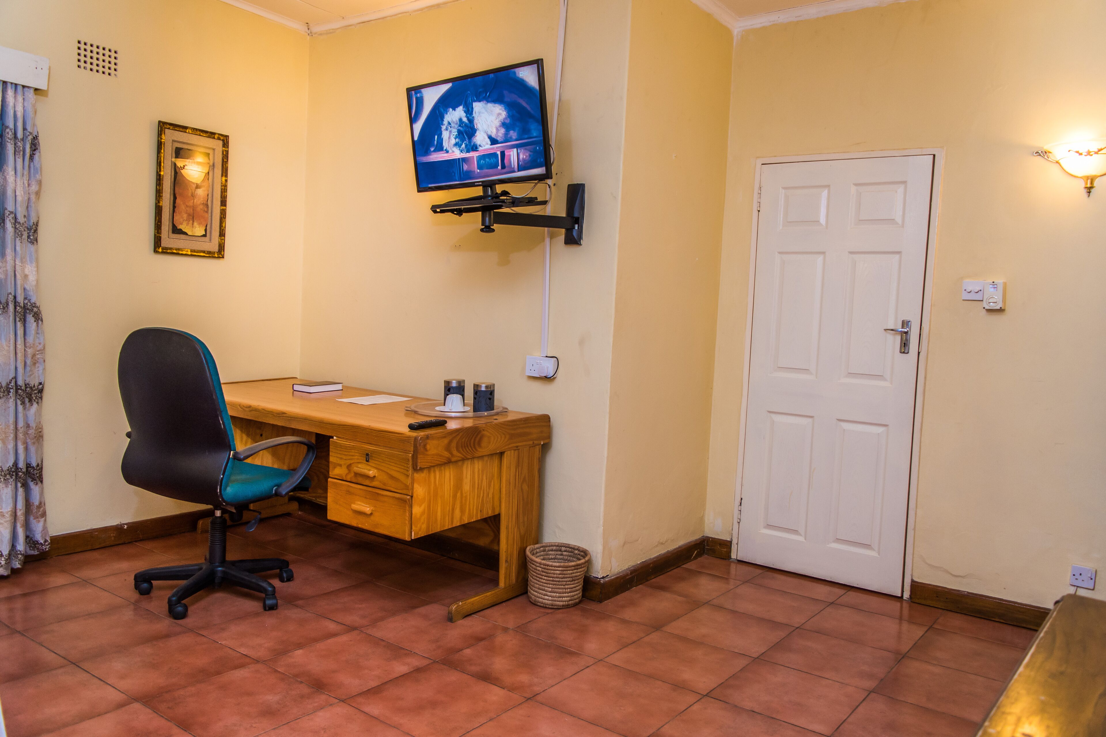 Een kamer met een houten bureau, een zwarte bureaustoel, een aan de muur bevestigde televisie, een witte deur en een kleine prullenbak.