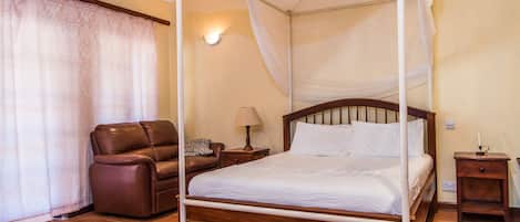 A four-poster canopy bed with a wooden frame and white bedding, a leather armchair, a bedside table with a lamp, and a wall-mounted light fixture.