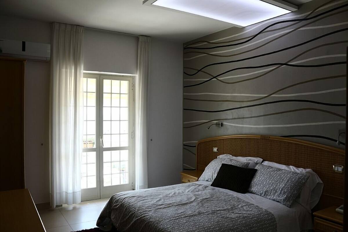 A bedroom with a bed, a nightstand, a window with curtains, and a wall with a wavy pattern.