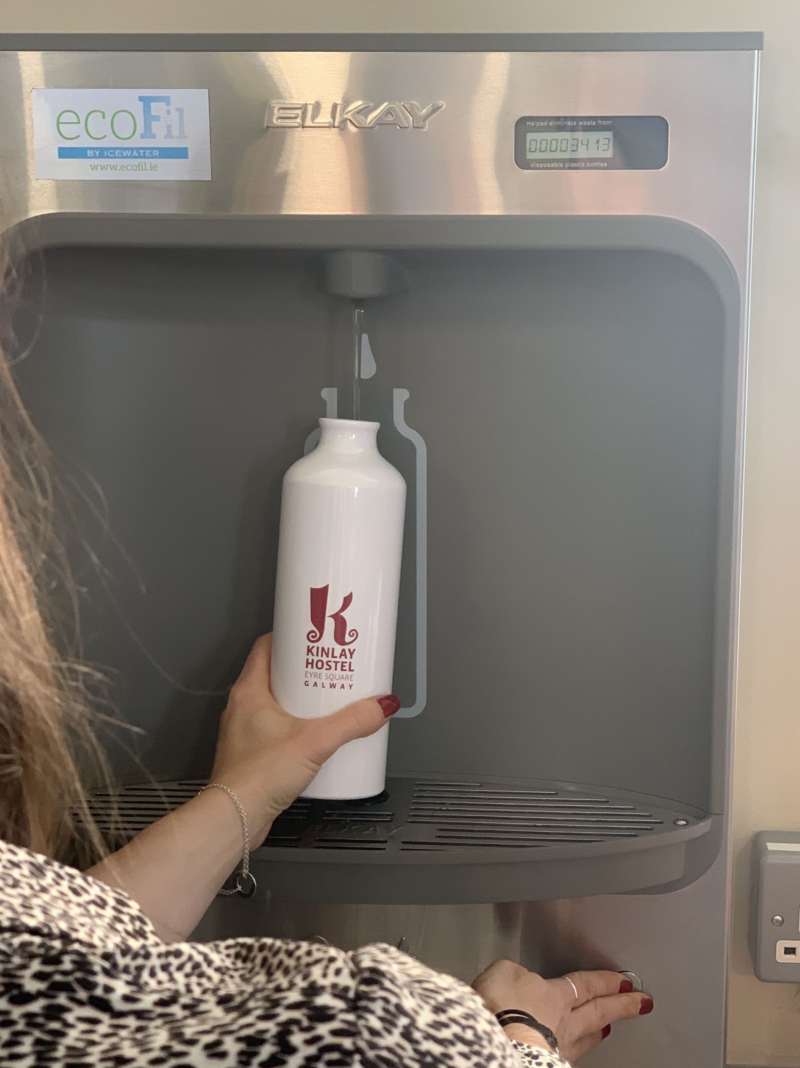 A water dispenser with a bottle of Kinlay Hostel water, a logo, and a recycling symbol.