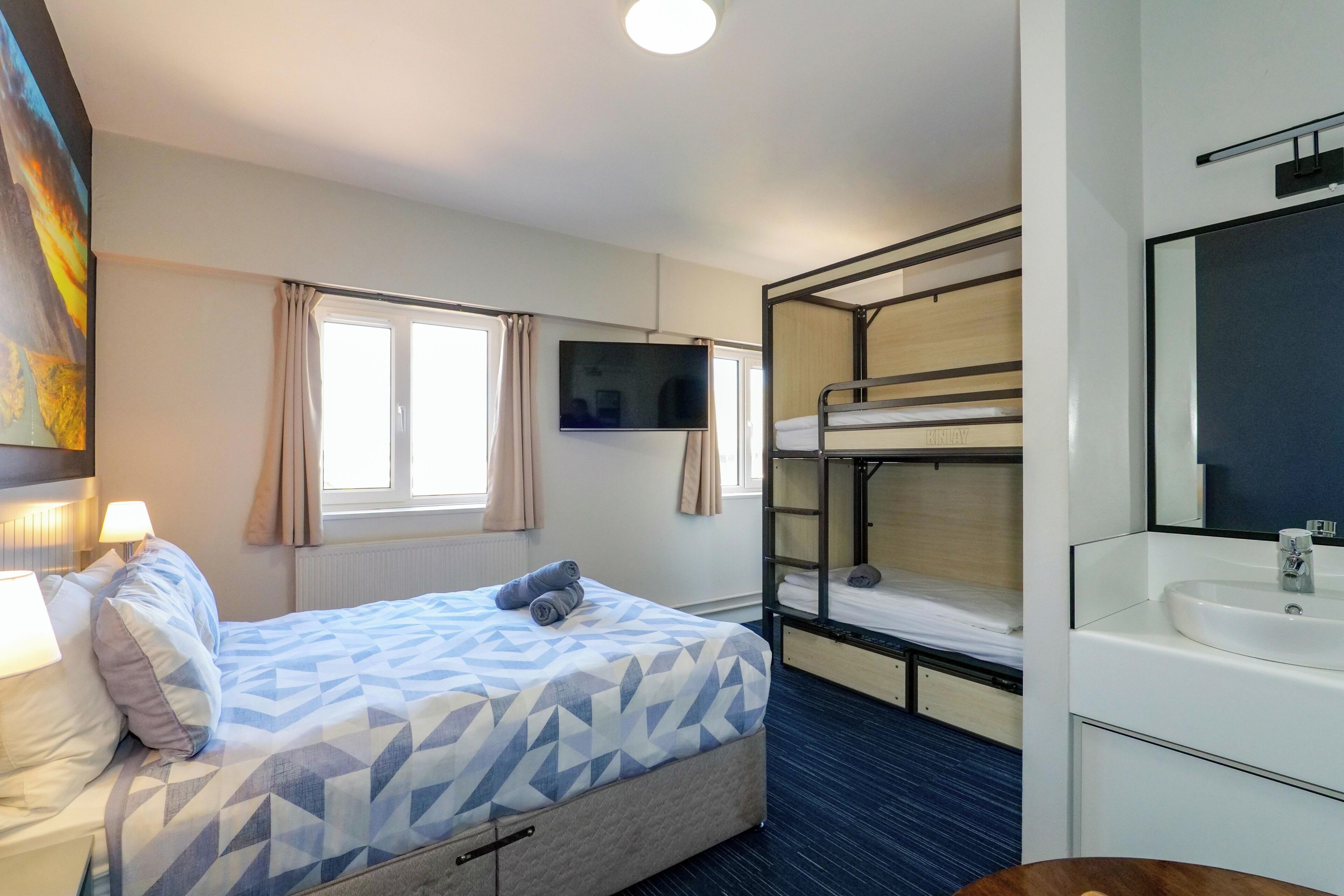 A hotel room with a bed, a TV, a bunk bed, a sink, and a window with curtains.