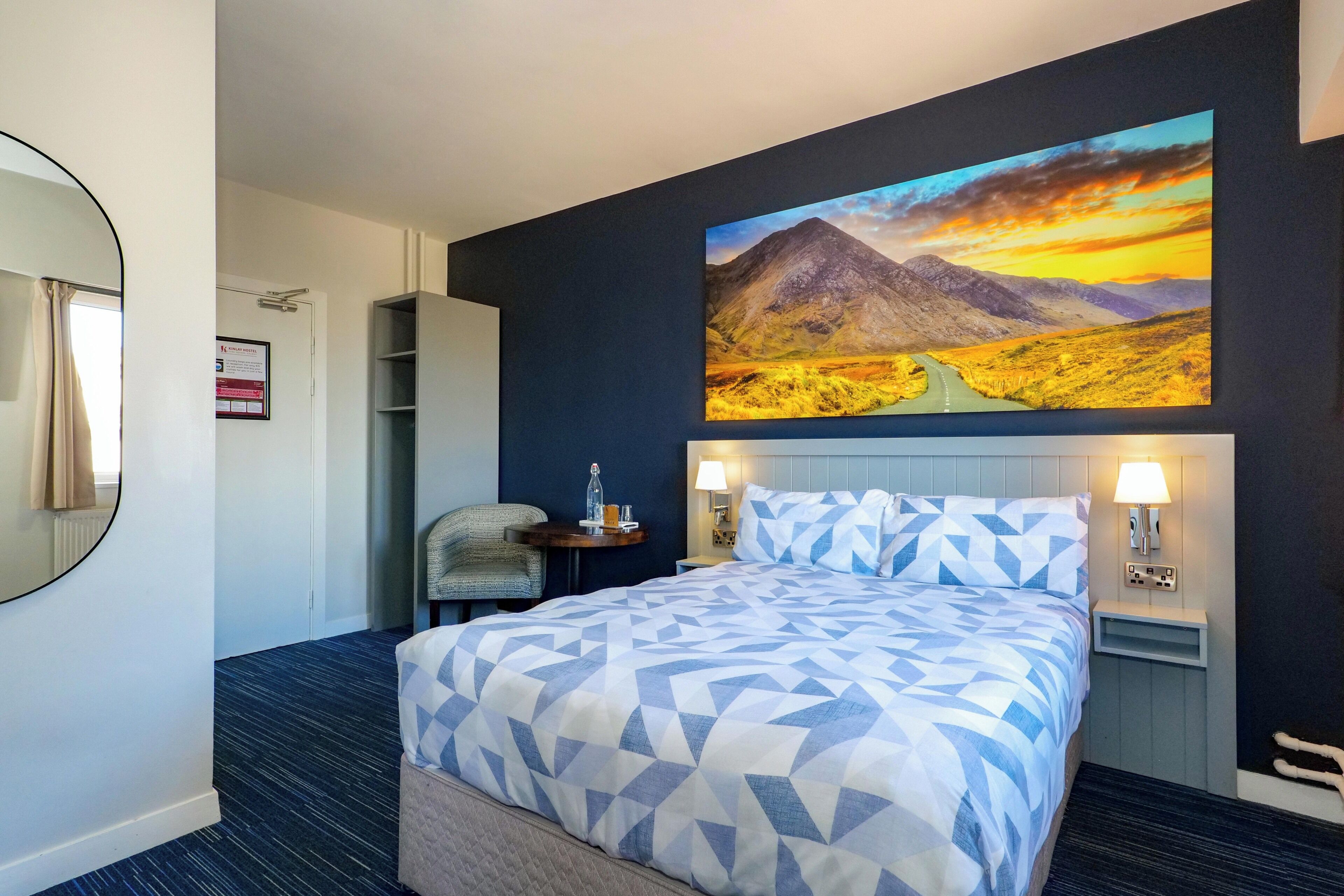 A hotel room with a bed, a nightstand with a lamp, a chair, a desk, a mirror, and a painting of a mountain landscape.