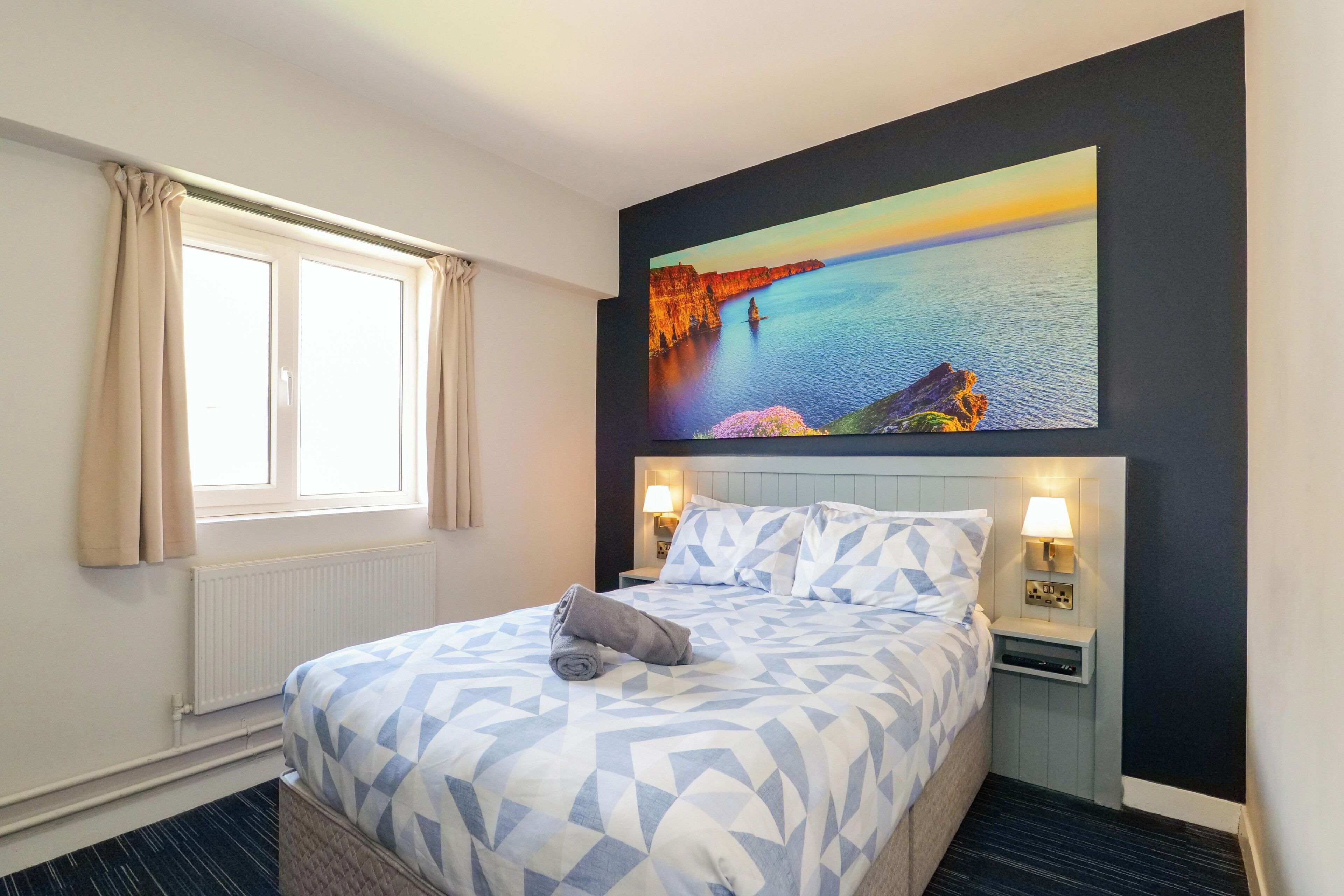 Quarto de hotel com cama, janela com cortinas, mesa de cabeceira com abajur e um quadro na parede.