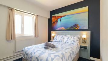 Quarto de hotel com cama, janela com cortinas, mesa de cabeceira com abajur e um quadro na parede.