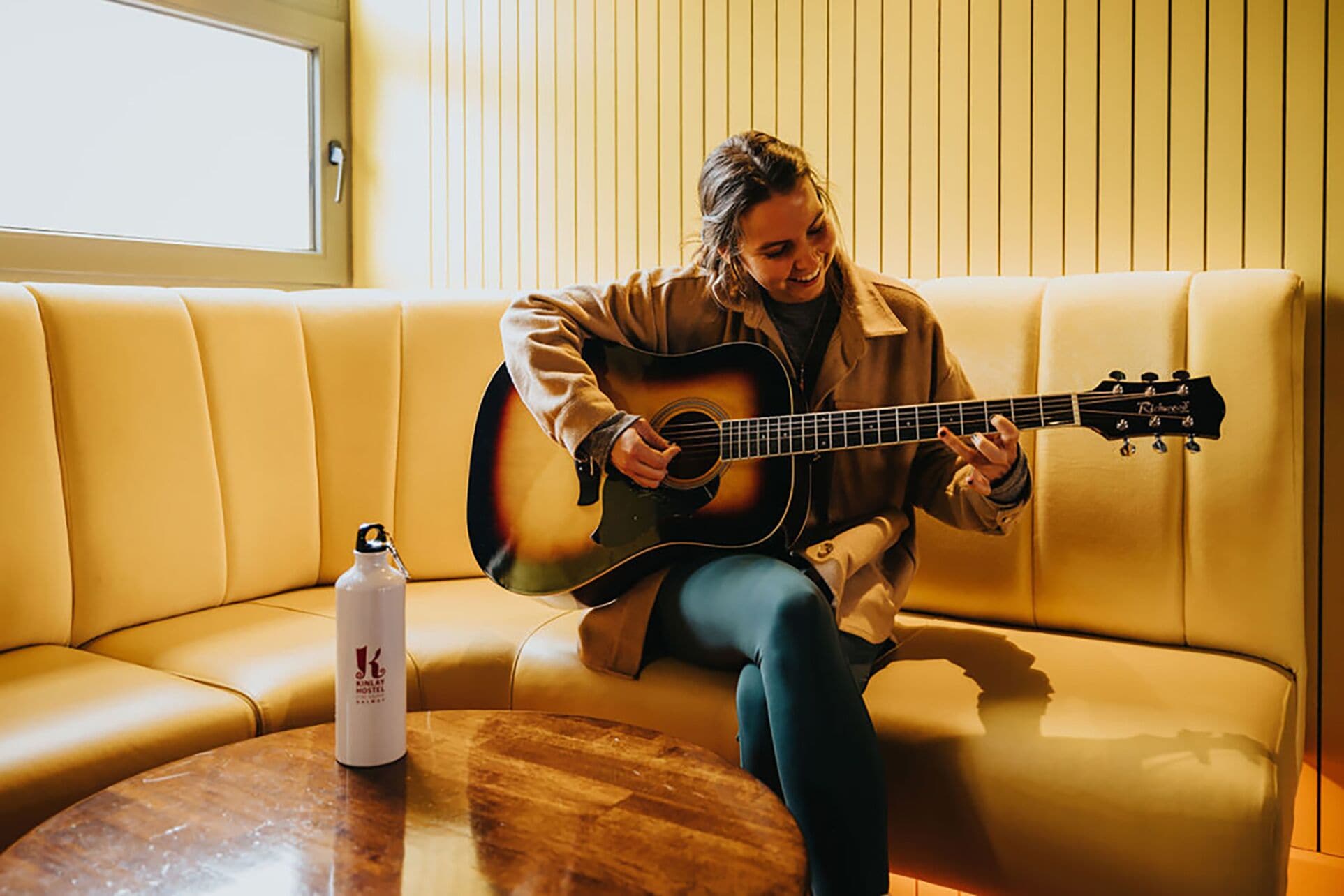 En person sidder på en beige sofa og spiller guitar. Der er en hvid flaske på et træbord ved siden af sofaen.
