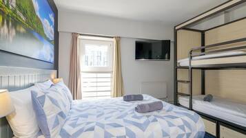 A hotel room with a bed, a TV, a window with curtains, and a bunk bed.