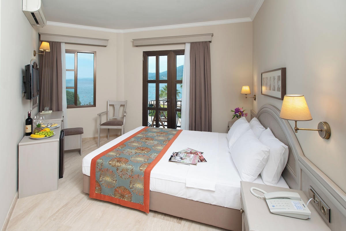 Ein Hotelzimmer mit Bett, Nachttisch, Lampe und Meerblick durch das Fenster.