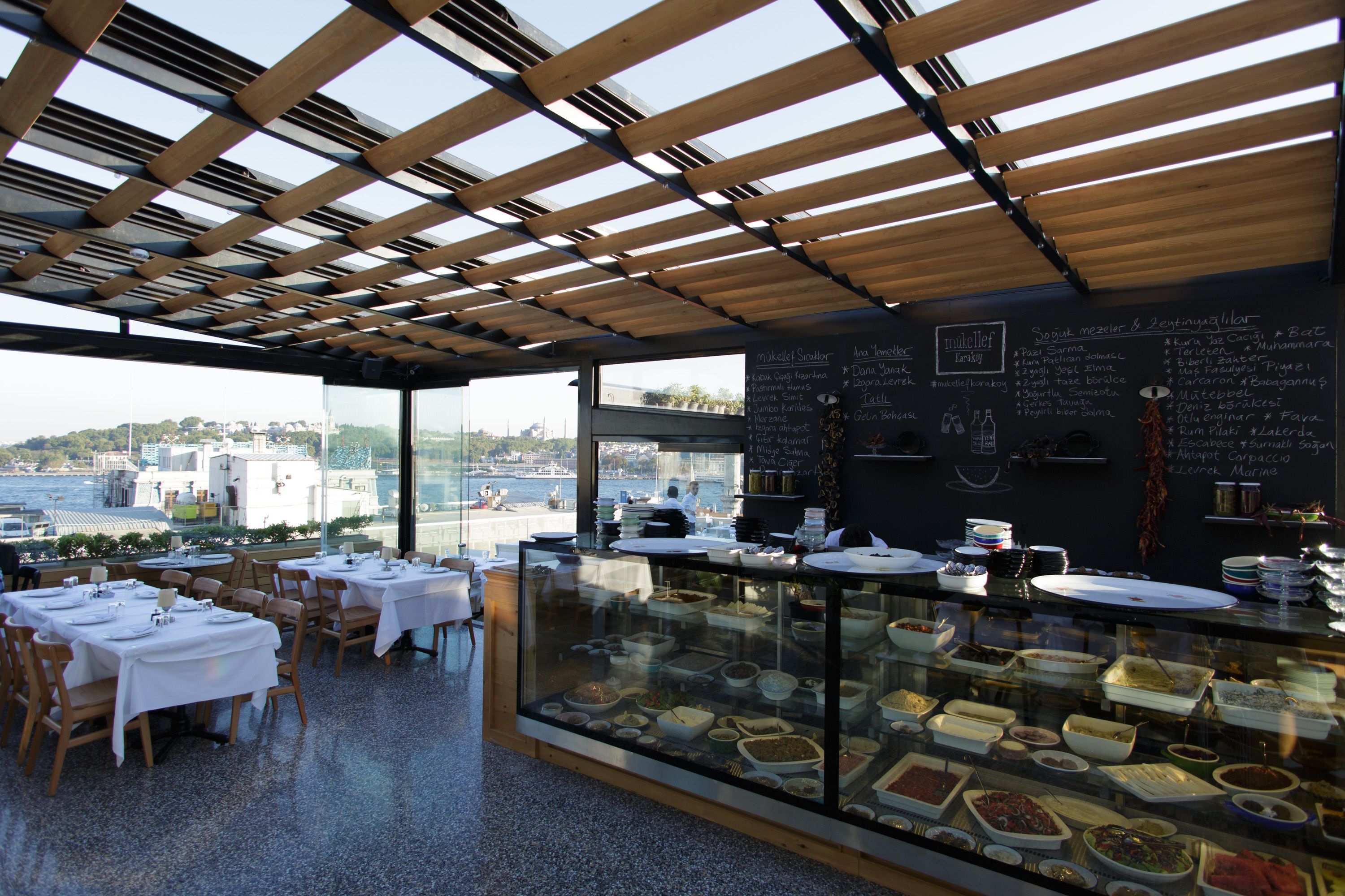 A restaurant with tables set for dining, a glass display case with food items, and a view of the city through large windows.