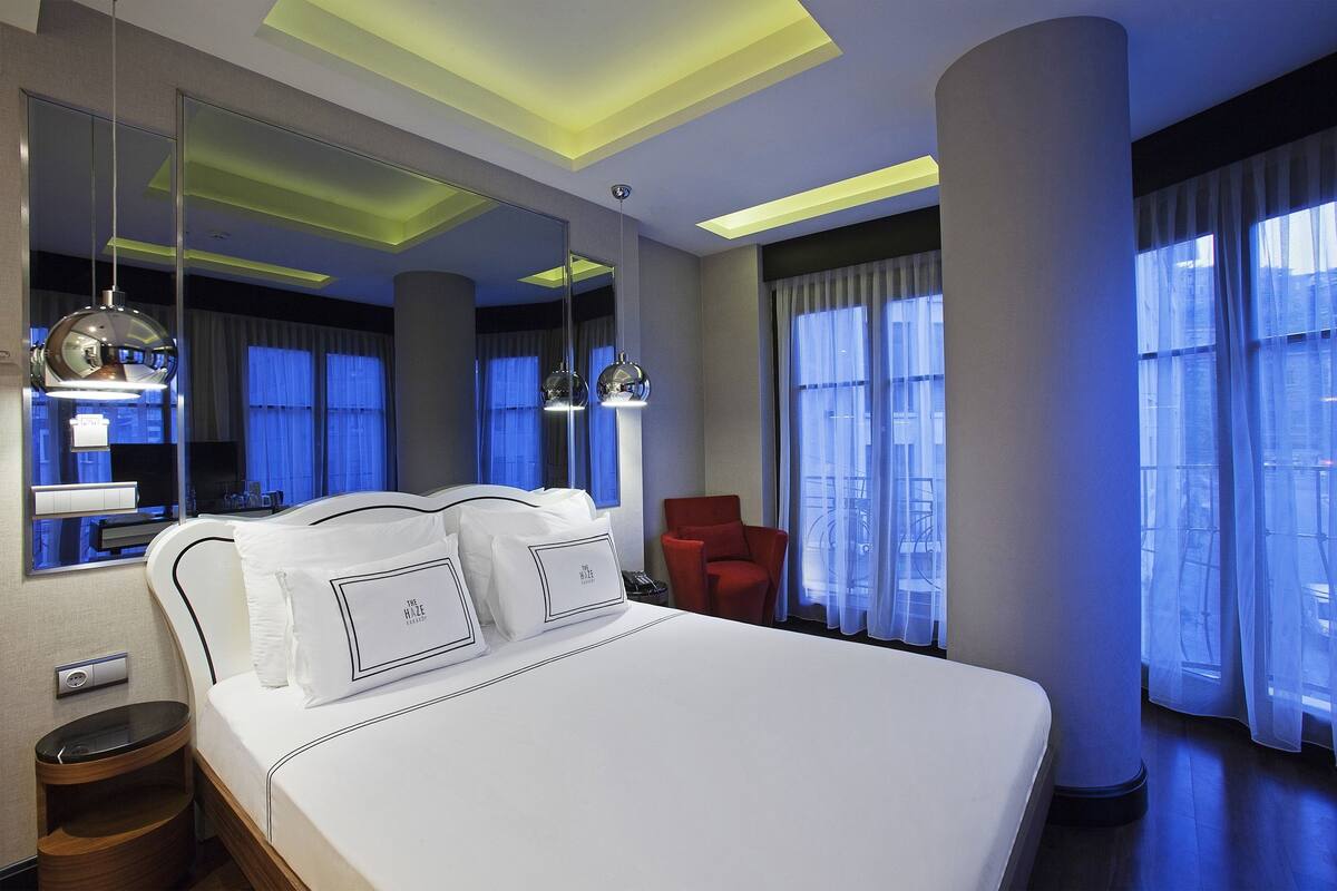A modern hotel room with a large bed, a red armchair, a large mirror, and a view of the city through the windows.