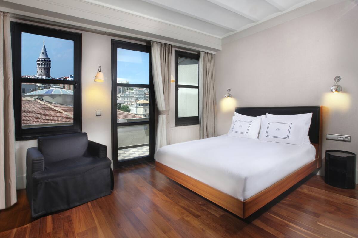 A hotel room with a large bed, a chair, and a view of a cityscape through a window.