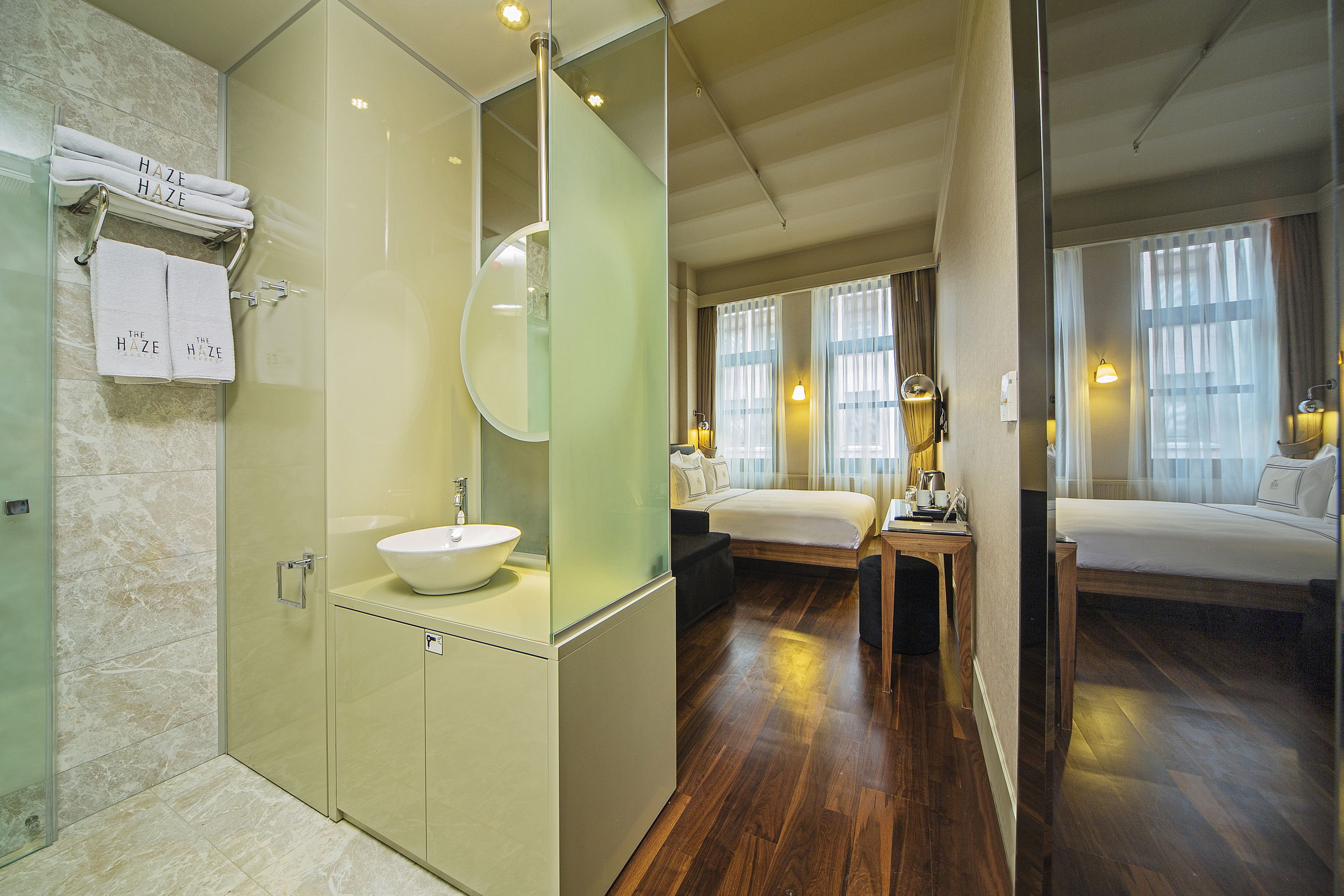 A modern hotel room with a glass-enclosed shower, a large mirror, a bedside table with a lamp, and a bed with a nightstand.
