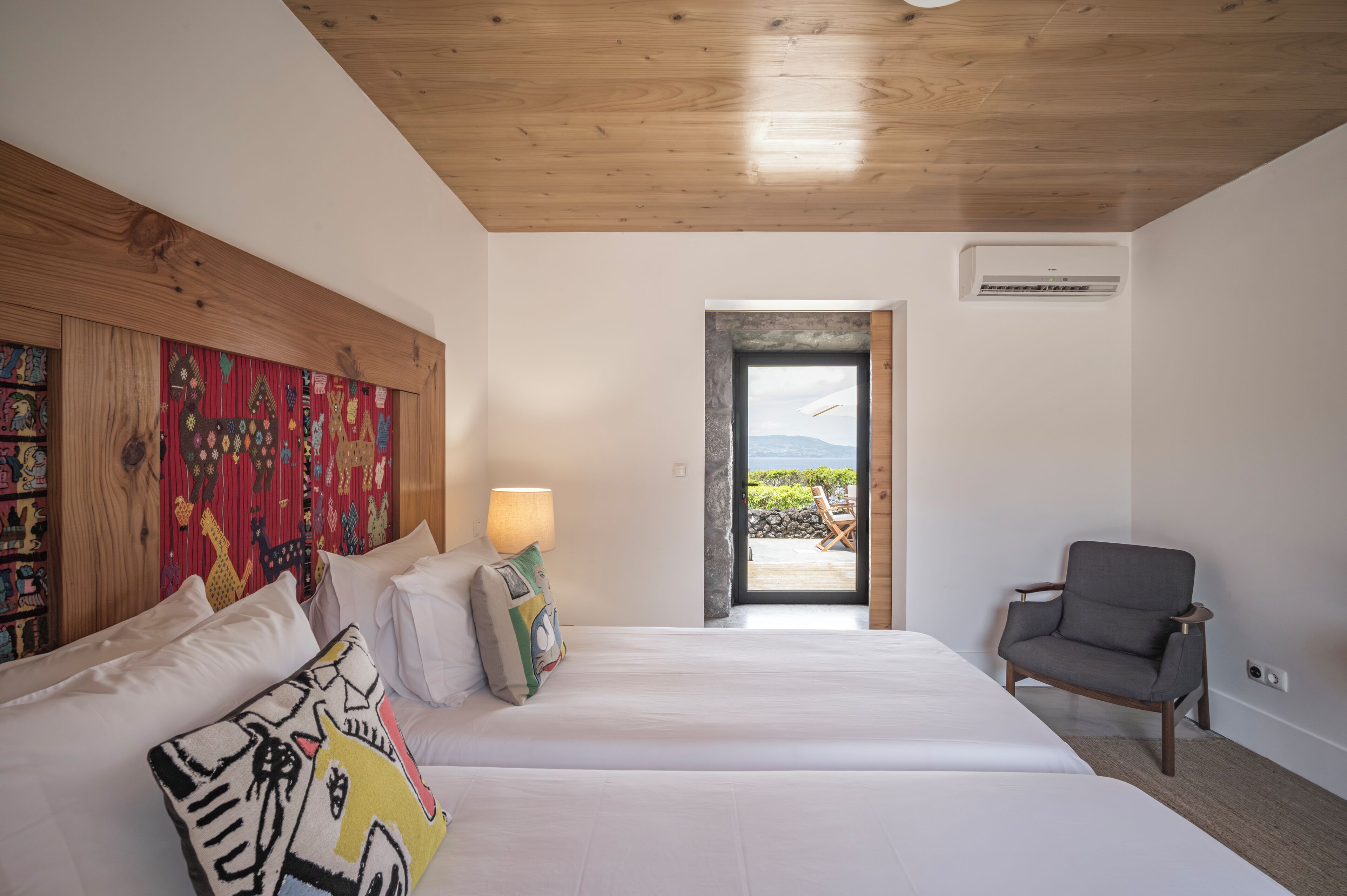 A hotel room with a wooden ceiling, a bed with white bedding, a chair, and a view of the outdoors.