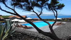 A pool area with lounge chairs and umbrellas, surrounded by rocky terrain and pine trees, overlooking a coastal landscape. - Pocinhobay (Madalena)