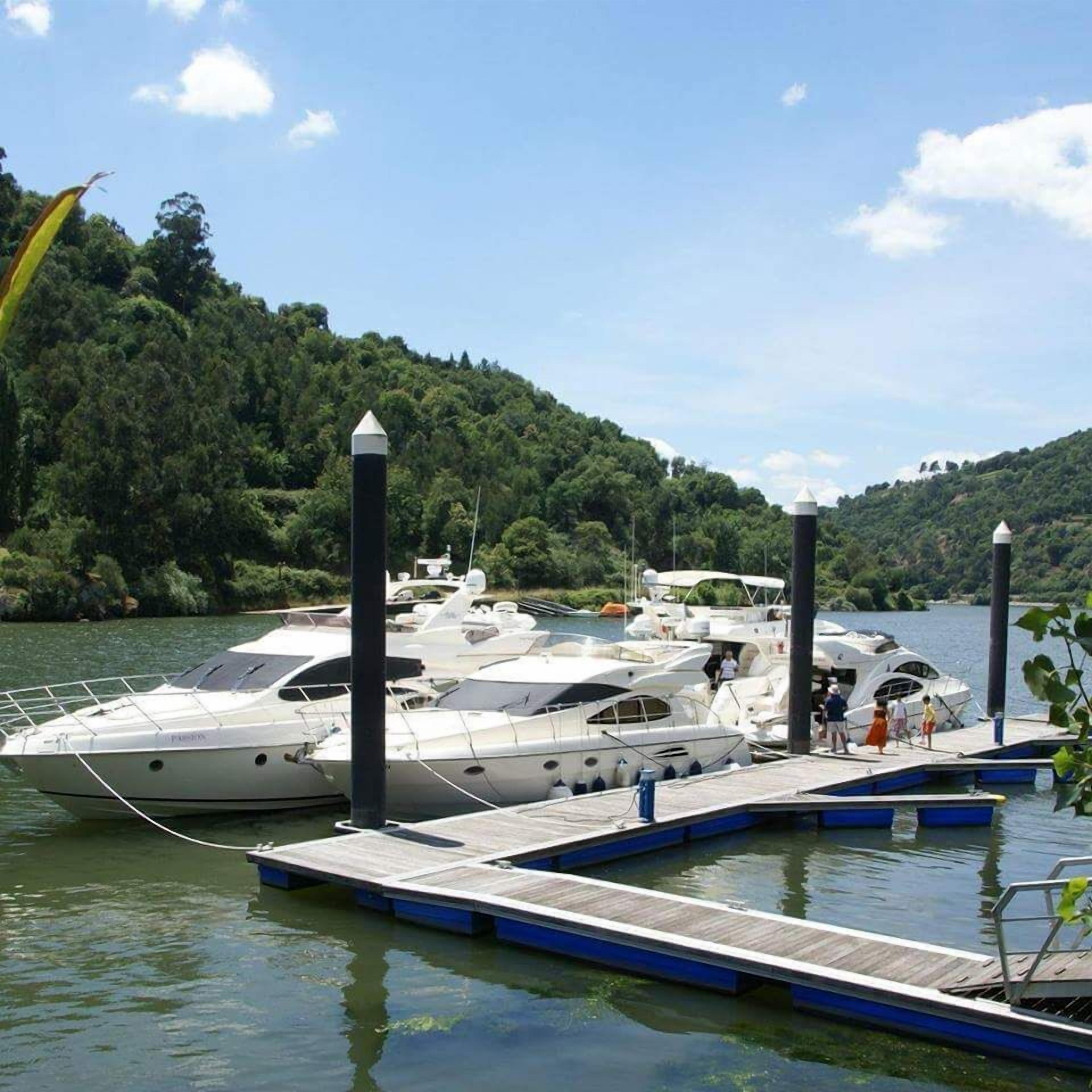 A marina with several yachts docked at wooden piers, surrounded by lush greenery and hills.