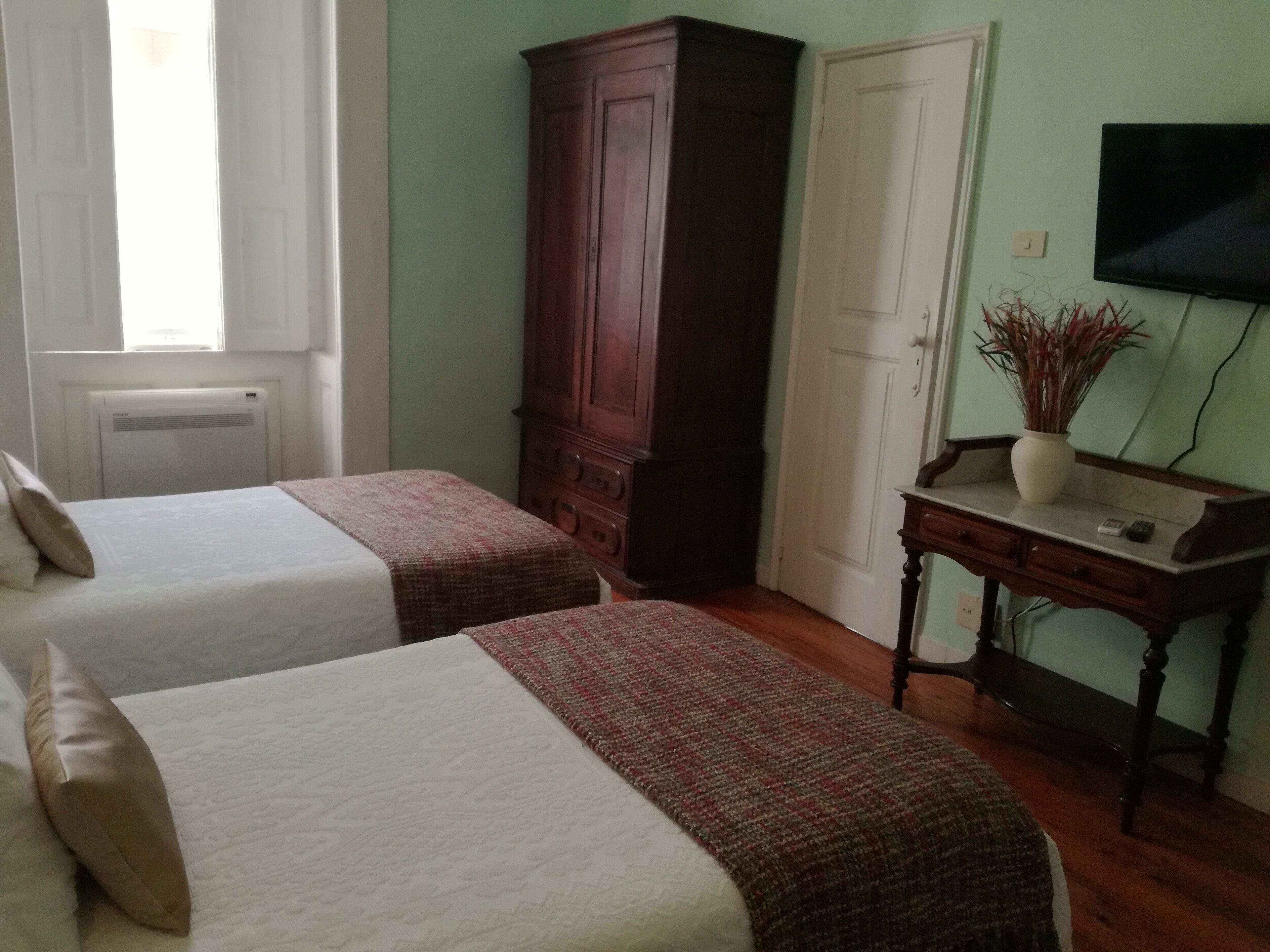 A room with two beds, a wooden wardrobe, a desk with a vase, and a television on the wall.