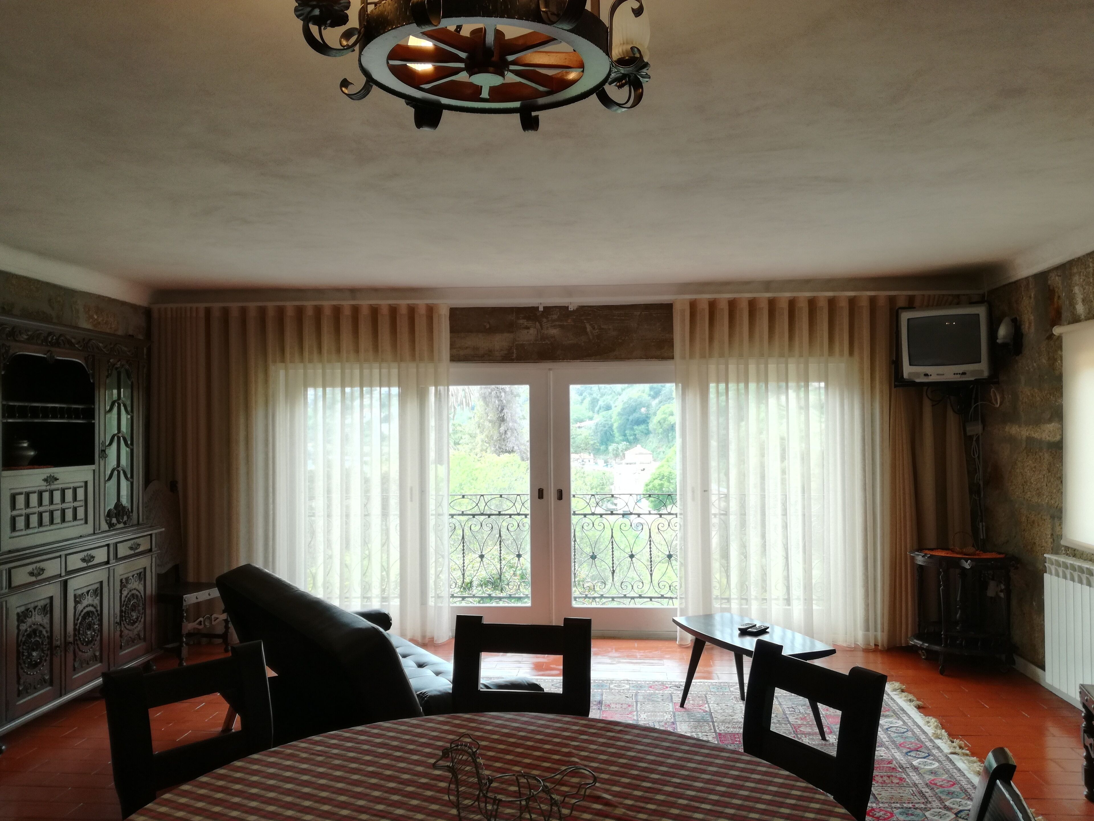A dining area with a round table, chairs, and a chandelier. There is a television mounted on the wall and a window with curtains.
