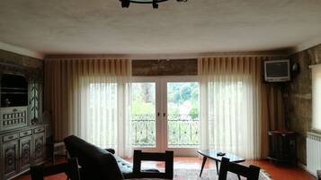 A dining area with a round table, chairs, and a chandelier. There is a television mounted on the wall and a window with curtains.