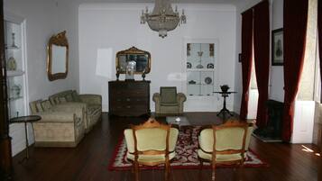 Een klassieke woonkamer met een kroonluchter, een zithoek, een eettafel met stoelen, een open haard en een spiegel aan de muur.