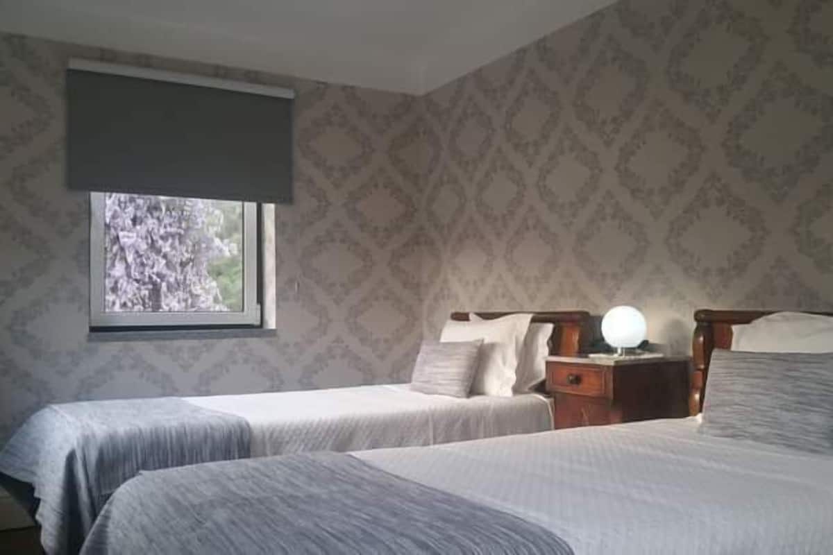 Une chambre avec deux lits, une table de chevet avec une lampe et une fenêtre donnant sur les arbres.