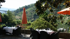 Un área de comedor al aire libre con mesas y sillas, rodeada de abundante vegetación y vistas a campos en terrazas.