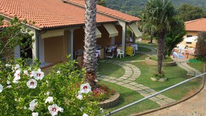 A villa with a tiled roof, a covered patio, and a landscaped garden with palm trees and flowering plants.