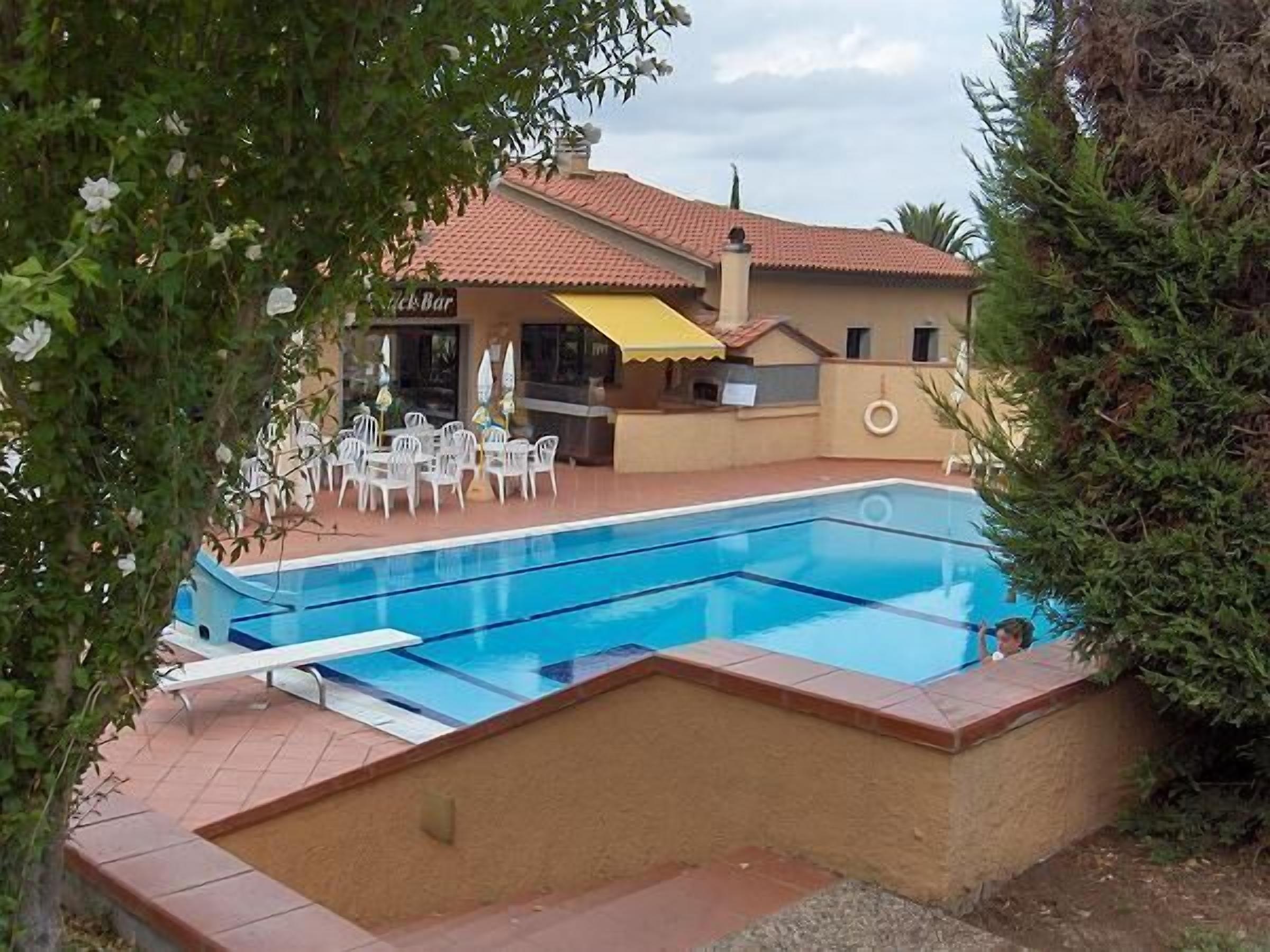 A swimming pool with a tiled deck, surrounded by a low wall and a building with a red-tiled roof.