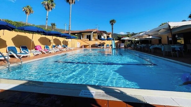 A swimming pool with lounge chairs and umbrellas, surrounded by palm trees and buildings.