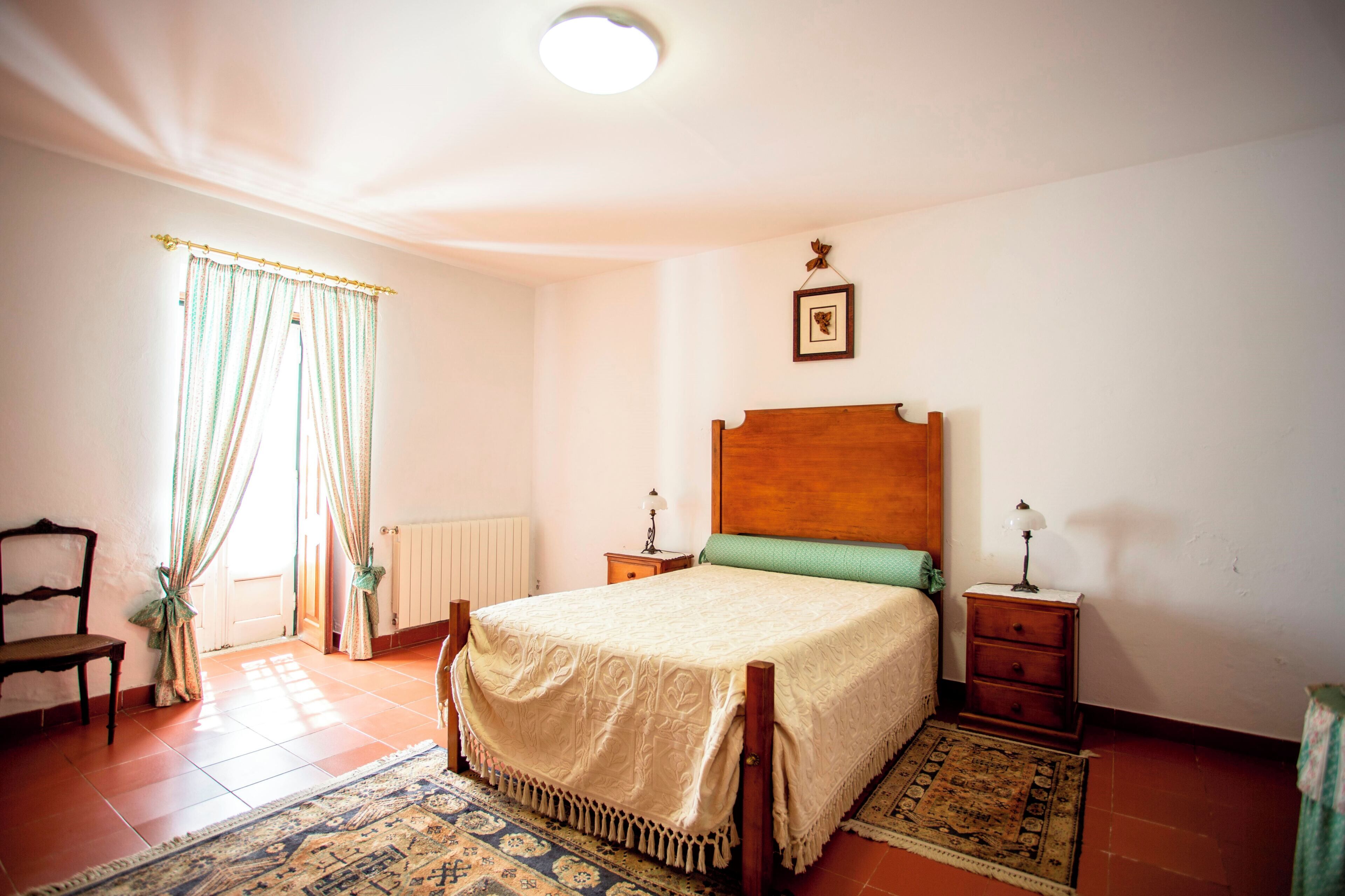 Un dormitorio con una cama de madera, dos mesitas de noche, una silla, un radiador y una ventana con cortinas.