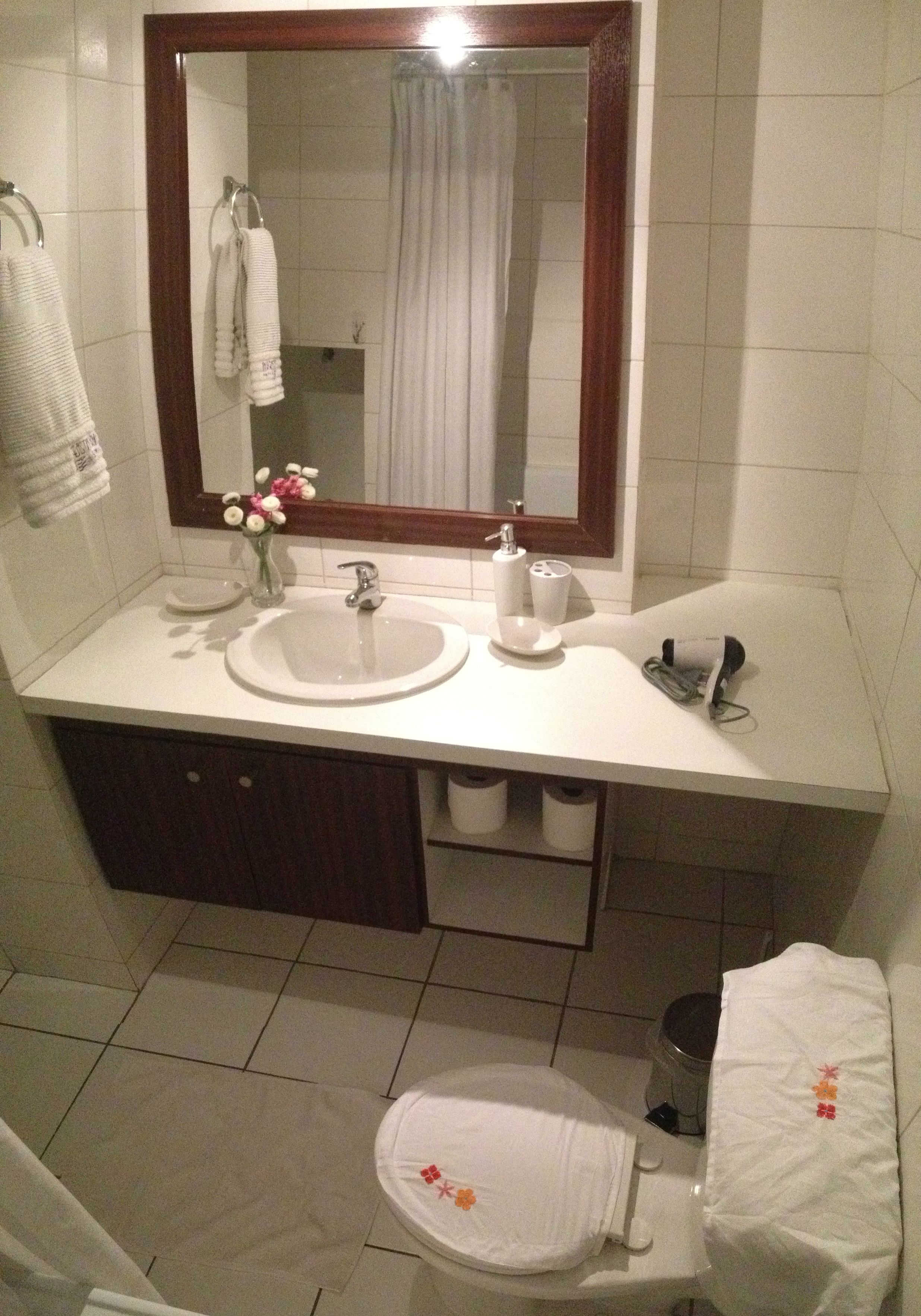 A bathroom with a white sink, mirror, and toilet. There is a towel rack with towels, a small vase with flowers, and a trash can.