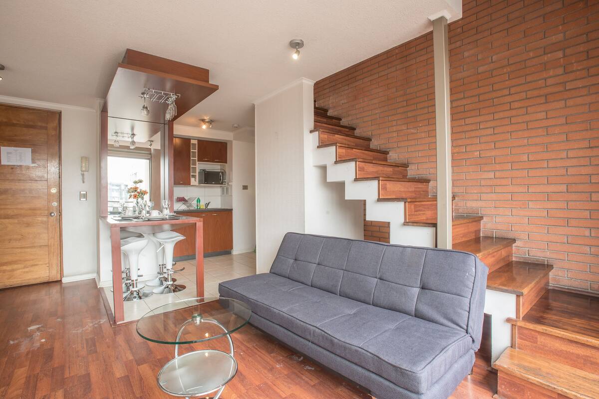 A modern living room with a grey sofa, a glass coffee table, and a kitchen in the background.