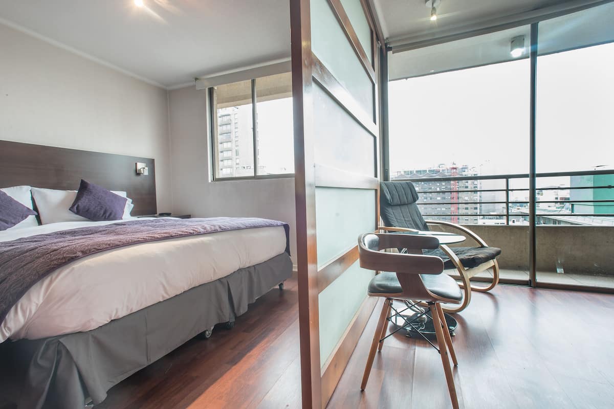 A bedroom with a large bed, a balcony with a view of the city, and a chair.