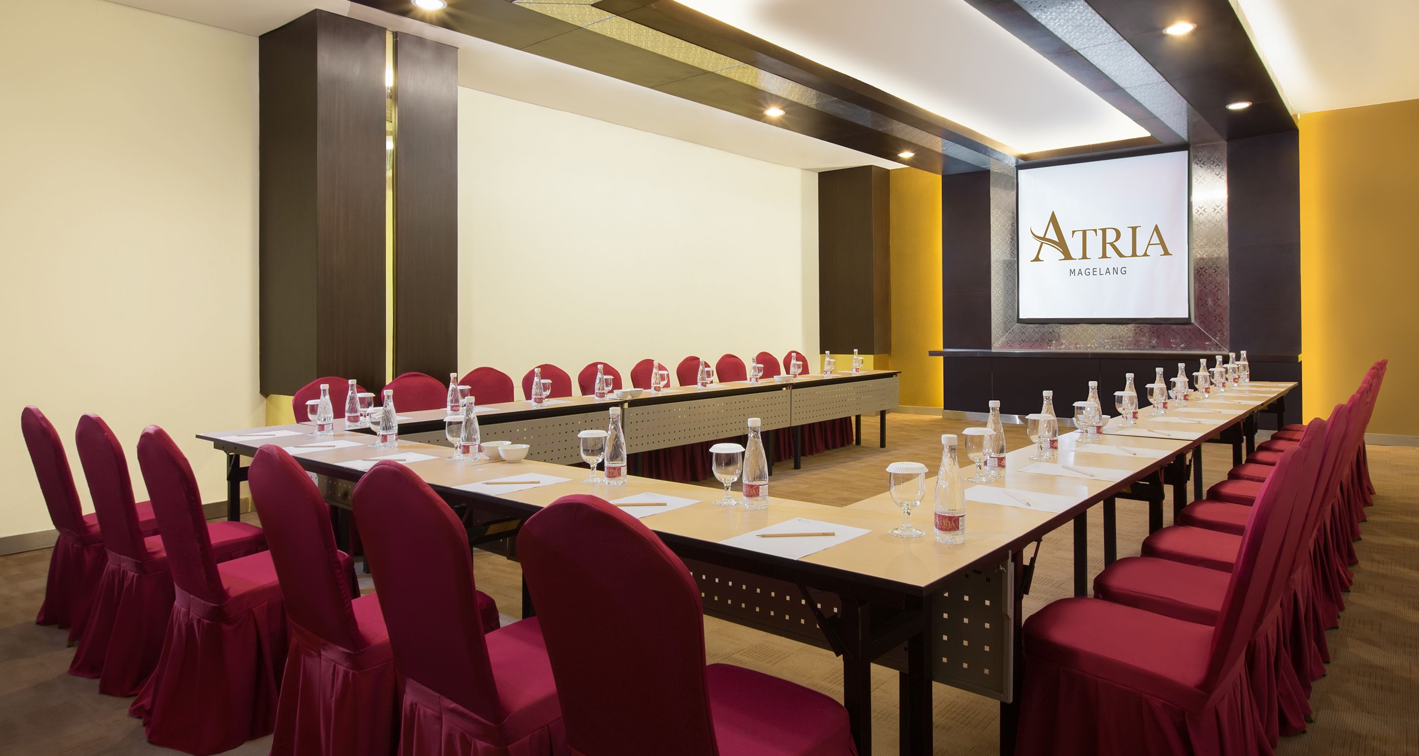 A conference room with a long table, red chairs, and a screen displaying "ATRIA MAGELANG".