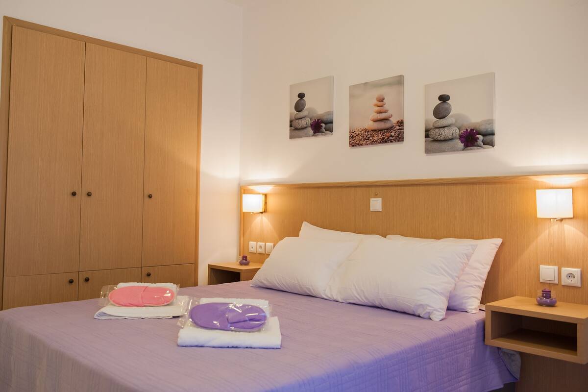 A hotel room with a large wardrobe, a bed with white and purple bedding, and three wall-mounted artworks depicting stacked stones.