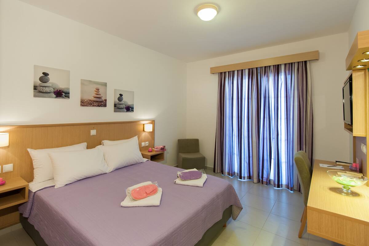 A hotel room with a bed, a desk, a chair, and a TV. There are three paintings on the wall.